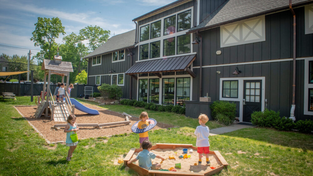 Watch "Franchising with Haven" - Our Childcare Franchise Opportunity 3 Children playing outside at Haven Gladstone Playground in New Jersey, featuring a wooden play structure, sandbox, and modern preschool building under a sunny blue sky.