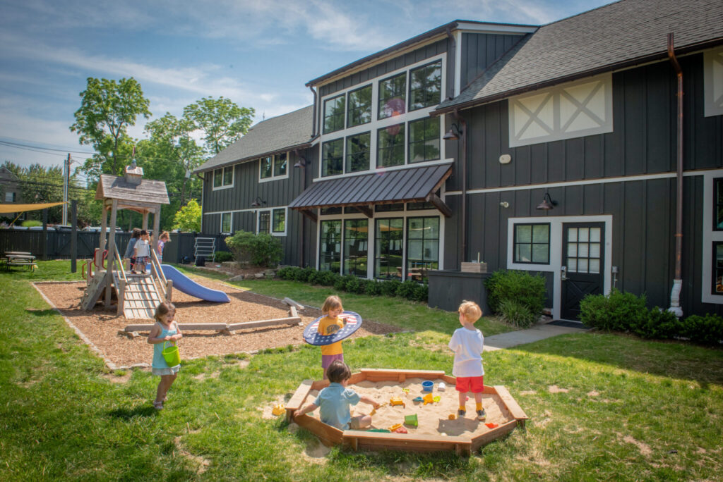 Watch "Franchising with Haven" - Our Childcare Franchise Opportunity 7 Children playing outside at Haven Gladstone Playground in New Jersey, featuring a wooden play structure, sandbox, and modern preschool building under a sunny blue sky.
