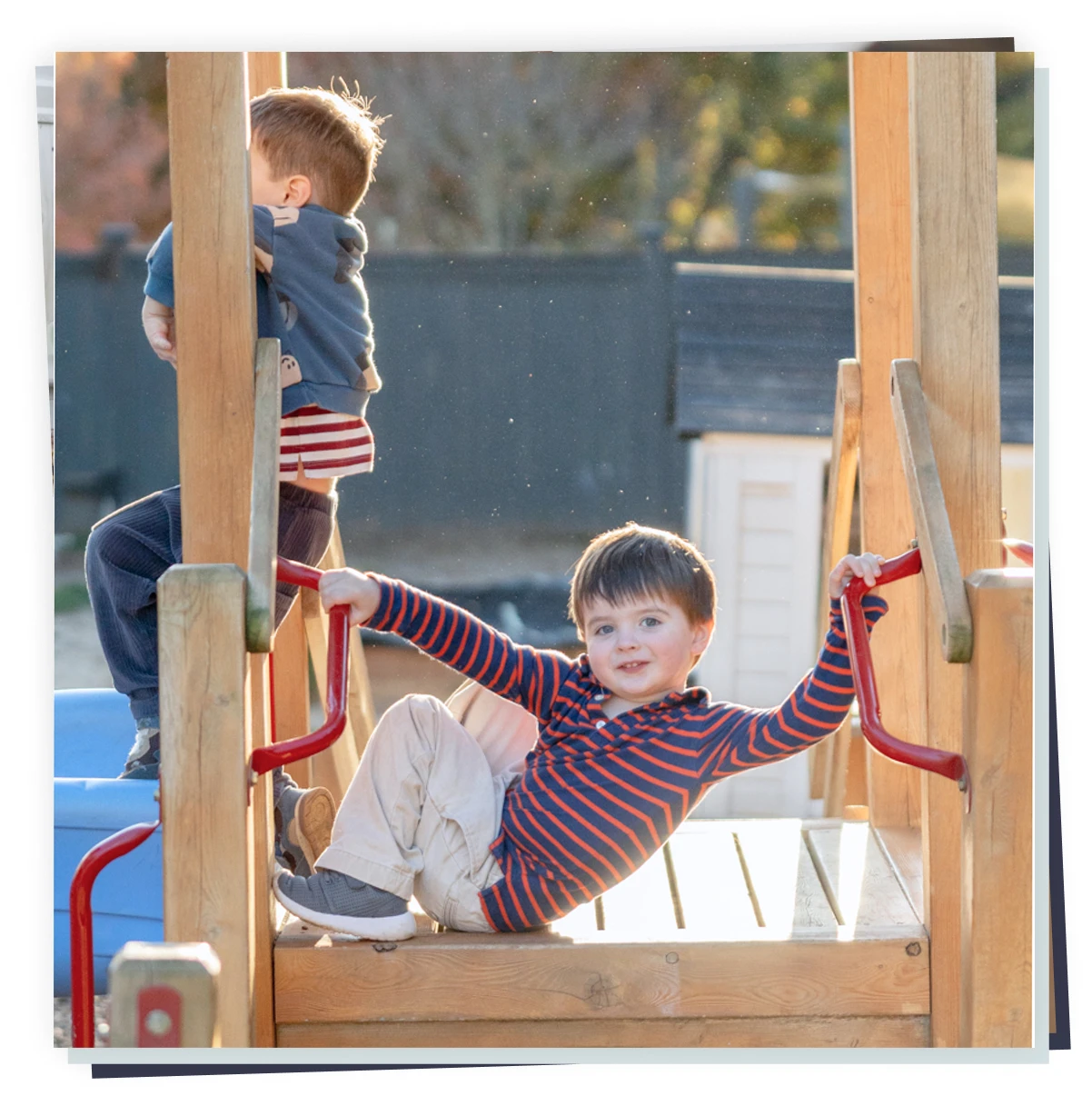 Two toddlers playing on a wooden playground platform, smiling child holds red rails, childcare outdoor play.