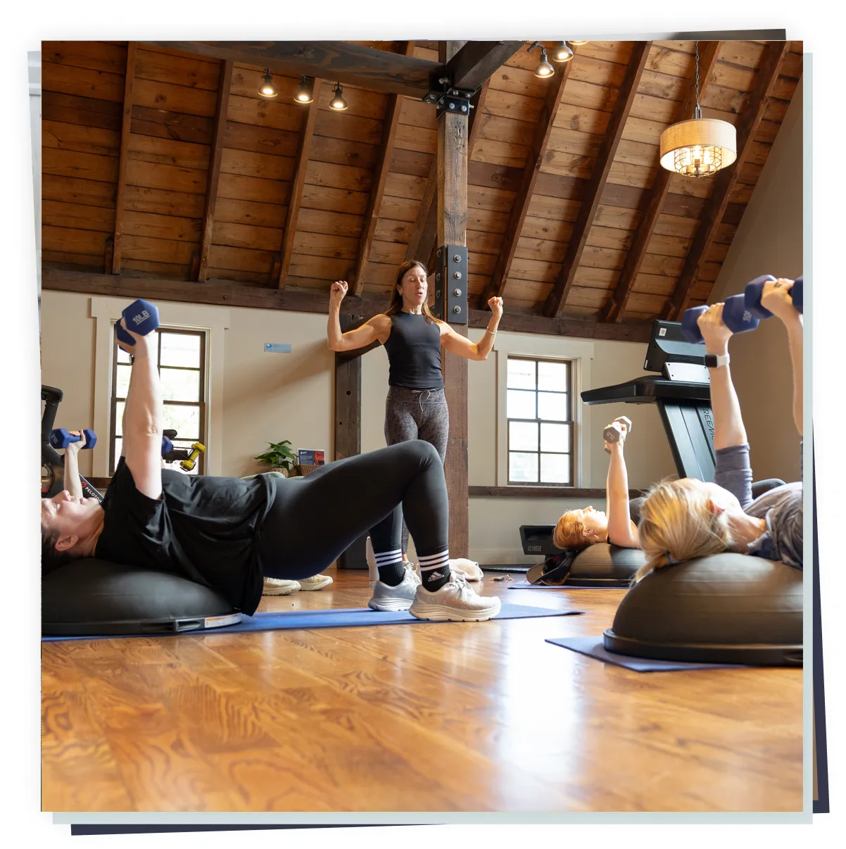 Instructor leading a fitness class on Bosu balance pads as participants lie down and lift dumbbells, wellness