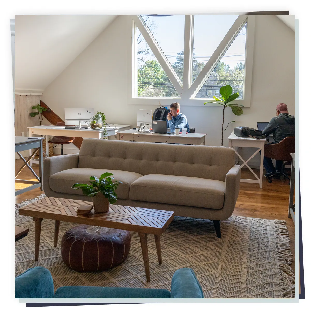 Sunlit coworking parent workspace with sofa, coffee table and two adults working at desks by large window