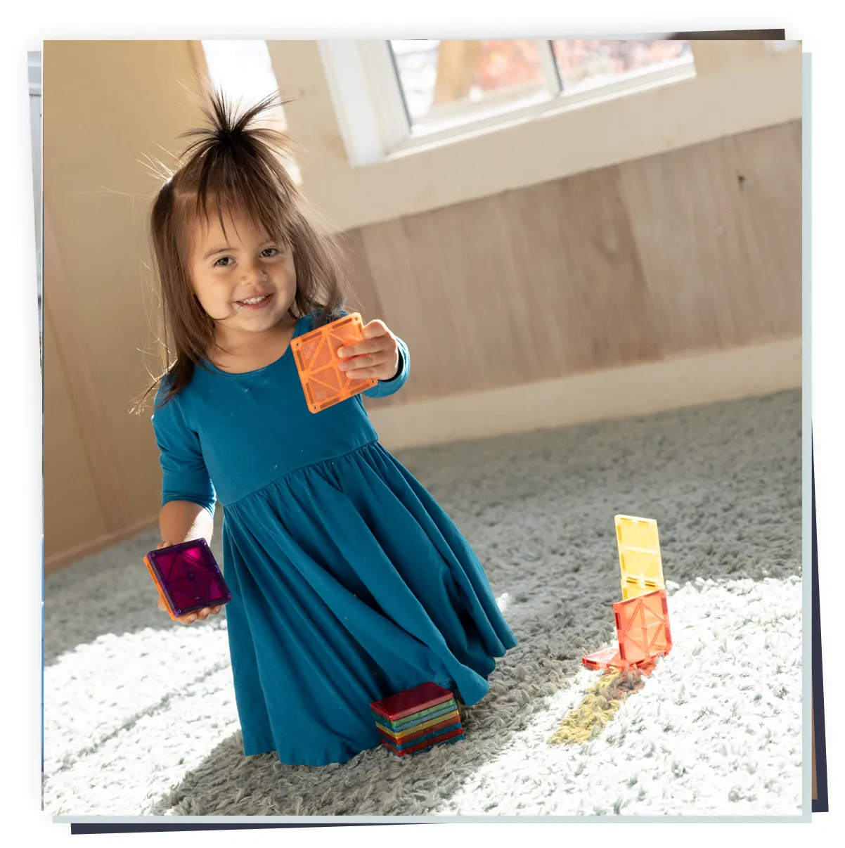 Toddler girl in blue dress kneeling on carpet, smiling while playing with colorful magnetic tiles; early learning play.