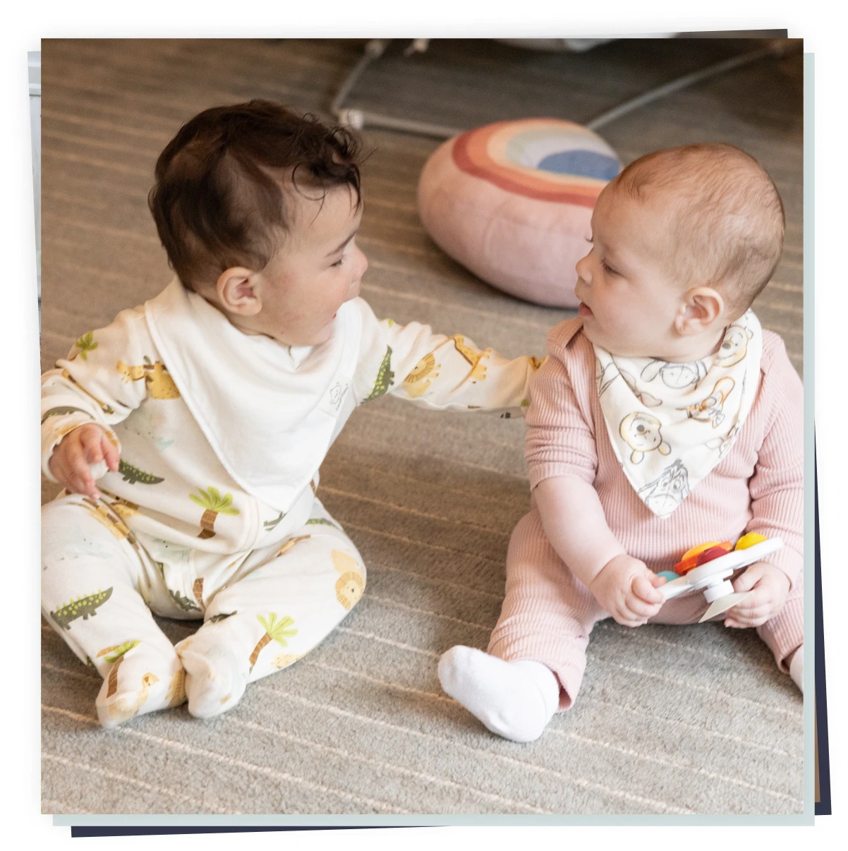 Two infants seated on a rug, one reaching to touch the other during play in a childcare setting.
