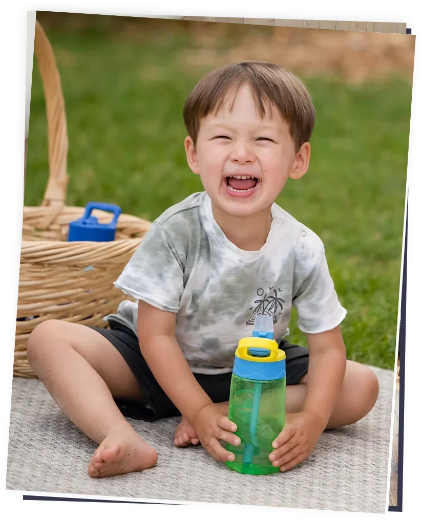 Laughing toddler seated on a blanket outdoors holding a green sipper bottle, picnic basket nearby, playful childcare moment.