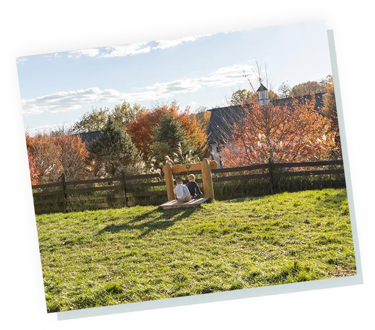 Two children sitting on a wooden play bench in a grassy yard with fence and autumn trees, Haven outdoor childcare.
