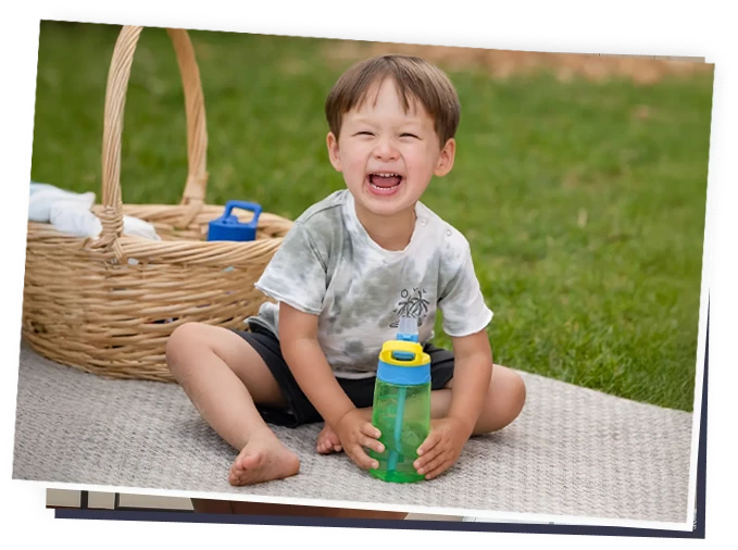 Smiling toddler sitting on an outdoor blanket holding a green sippy cup, playful childcare scene